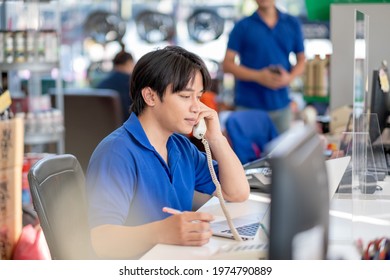 Asian Man Receive Phone Call From Customer And Stay In Counter Service Area Of Automotive Or Garage Shop With Other Worker Work In The Background.