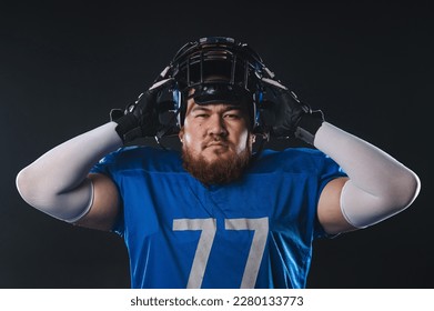 An Asian man with a red beard in a blue american football uniform puts on a helmet on a black background. - Powered by Shutterstock