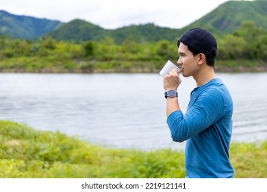 Asian Man Relax And Enjoy Outdoor Lifestyle Hiking And Camping In Forest Mountain While Travel On Summer Holiday Vacation Trip. Handsome Guy Brewing And Drinking Hot Coffee By The Lake In Morning.