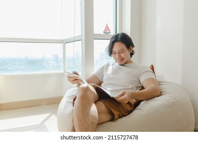 Asian Man Relax And Reading Book On Bean Bag Sofa In His Apartment.