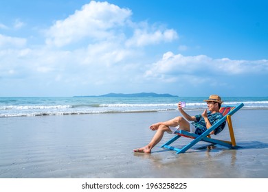 Asian Man Resting On Sunbed On The Beach. Happy Guy Sitting On Beach Chair By The Sea Using Smartphone Selfie Or Video Call With Friend. Male Relax And Enjoy Beach Outdoor Lifestyle On Summer Vacation