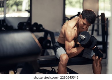 Asian man shirtless workout weight training biceps muscles with dumbbell in fitness gym. Weight training exercise in concept of health and wellness.