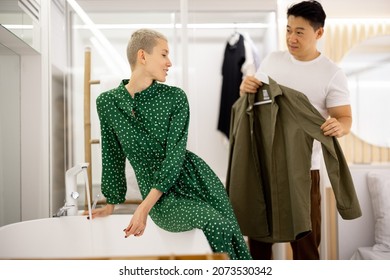 Asian Man Showing Blazer To His Caucasian Wife. Couple Getting Ready To Spending Time Together In Public Place. Concept Of Family Relationship. Girl Sit On Bath With Pouring Water. Bathroom Interior