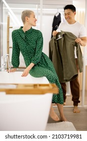 Asian Man Showing Blazer To His Caucasian Wife. Couple Getting Ready To Spending Time Together In Public Place. Concept Of Family Relationship. Girl Sit On Bath With Pouring Water. Bathroom Interior