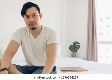 The Asian man sits in the bedroom with a laptop on the bed.