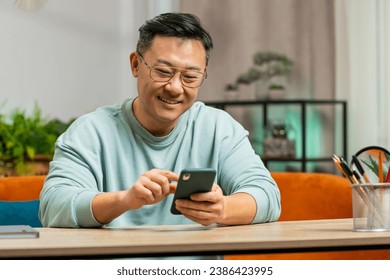 Asian man sits on couch uses mobile phone smiles at home room apartment. Middle-aged Chinese guy texting share messages content on smartphone social media applications online watching relax movie - Powered by Shutterstock