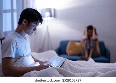 Asian man sitting in bed using a laptop, while a woman in the background appears upset and stressed, highlighting a tense moment in a relationship at night home - Powered by Shutterstock