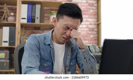 Asian Man Sitting Behind A Laptop Is Touching And Blinking His Eyes While Suffering Eye Irritation At Work In The Office.