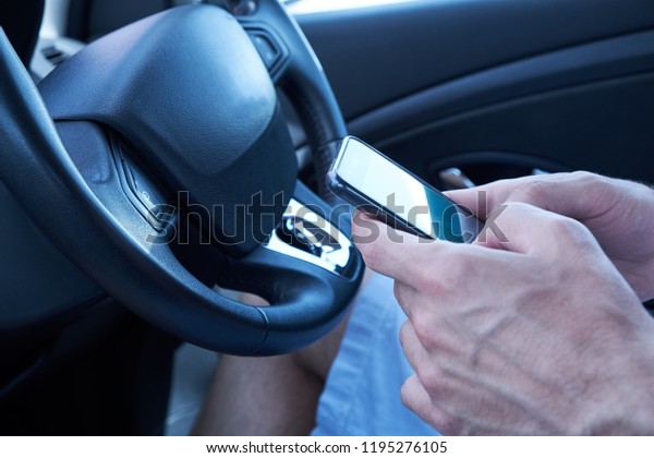 Asian man sitting in car with mobile phone in\
hand texting while\
driving