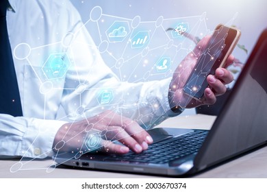 Asian Man Is Sitting At A Desk With His Left Hand Holding A Mobile Phone.  It Has A Hologram Showing Business Icons In The Hitech Circle. Background Blur