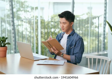 Asian Man Sitting In Front Of Laptop And Writing Notes