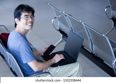 Asian Man Sitting On A Chair In A Waiting Room, Using Laptop And Smartphone While Listening Music With His Headphones. Looking Happily At The Camera.