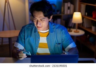 Asian Man Sitting On The Floor Is Holding Credit Card For Shopping Online By Laptop At Home