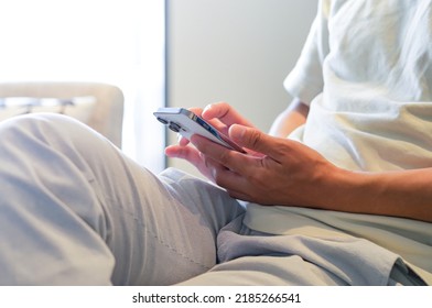 An Asian Man Sitting On The Sofa In The Living Room And Operating His Smartphone