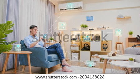 asian man sitting on sofa and using smart home control app on mobile phone with augmented reality view of IOT connected objects in apartment