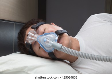 Asian Man With Sleep Apnea Using CPAP Machine, Wearing Headgear Mask Connecting To Air Tube