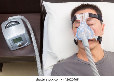 Asian man with sleep apnea using CPAP machine, wearing headgear mask connecting to air tube, selective focus on the man and headgear mask