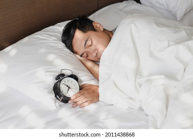 Asian Man sleeping on the bed under the blanket with alarm clock showing 7 oclock 