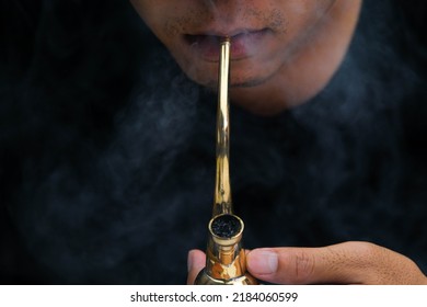 Asian Man Smokes Marijuana From A Pipe At Home. Studio Shoot With Model Simulating Smoking Pot With A Pipe In A Dark Background. Cannabis Legalisation.