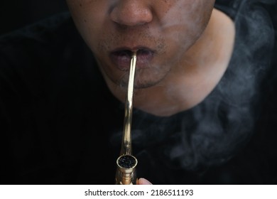 Asian Man Smokes Marijuana From A Pipe At Home. Studio Shoot With Model Simulating Smoking Pot With A Pipe In A Dark Background. Cannabis Legalisation.