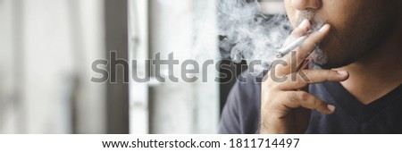 asian man smoking a cigarette stand by the window dark background. concept healthcare. banner copy space.
