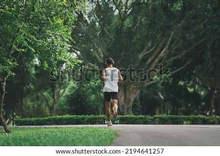 Asian man sportsman fit body wearing white headphone and smart watch while running or jogging in the garden have more tree and clean air, exercising at park. Sport healthy running concept.