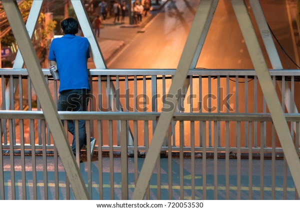 Asian man standing behind and\
holding a beer can on the footbridge and looking at the empty\
street not running cars, This is night time in the city of\
Bangkok.