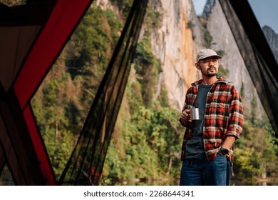 Asian man standing in front of a tent holding a hot drink mug. coffee and camping. - Powered by Shutterstock