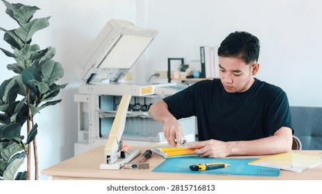 Asian man stapling a stack of yellow document covers at a work table in a copy shop with a large printer in the background.