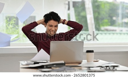 An Asian man struggles with workload, hands on head, surrounded by clutter and chaos in the office.