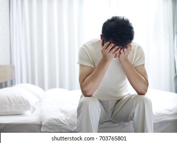 asian man suffering from insomnia sitting on bed head down covering face with hands.