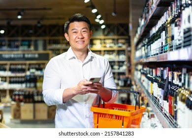 Asian Man In Supermarket, Uses Phone And App To Buy And Choose Wine In Store, Looks At Camera And Smiles