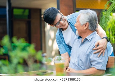 Asian man surprise hugging elderly father from back at outdoor garden cafe restaurant on summer vacation. Family relationship, holiday celebrating, father's day and old people health care concept