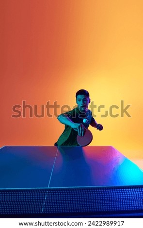 Asian man, table tennis player poised to strike in neon light against warm yellow gradient background. Dynamic gel portrait. Concept of professional sport, healthy lifestyle and recreation. Ad