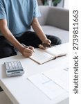 Asian Man Taking Notes on Finances, Budget, Household Expenses, and Tax Calculations in Notebook at Home