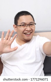 Asian Man Taking Selfie Photo Or Doing A Video Call At Home, Smiling To Camera And Waving. Happy Cheerful Expression