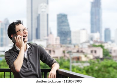 Asian Man Talking On Smartphone Smiling Laughing Out Loud Relaxing At Balcony With City View Copy Space Background. Candid Of Casual Lifestyle Freelancer Make Phone Call With Confident And Positive.