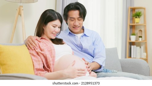 asian man is touching pregnant wife belly and expecting baby on couch at home