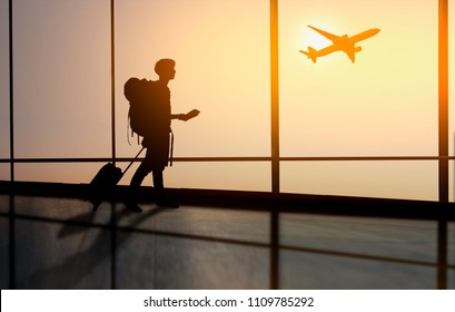 Asian Man Tourist Is Standing In Airport And Looking At Aircraft Flight Through Window. Tourists Hold A Ticket In Hand With Red Luggage At The Airport.