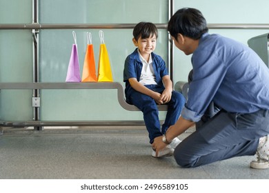 An Asian man is tying his son's shoelaces as the boy sits on a bench while they wait for the train at the railway station. - Powered by Shutterstock