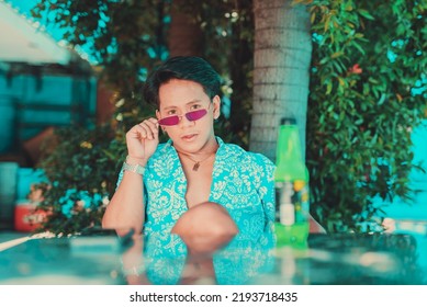 Asian Man In An Unbuttoned Hawaiian Luau Polo Shirt And Necklace Holding His Purple Sunglasses Near A Reflective Glass Table In A Garden.