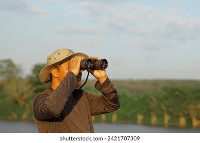 Asian man uses binoculars to observe local birds along a river. Soft and selective focus.                                 - Powered by Shutterstock