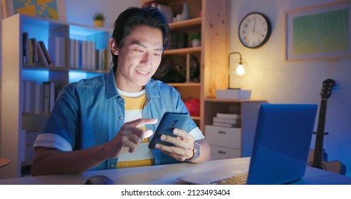 Asian Man Uses Phone And Laptop At Night In His Room