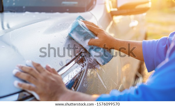 Asian\
man using blue sponge with soap to washing the car at outdoor in\
sunset time. Car cleaning and maintenance\
concept