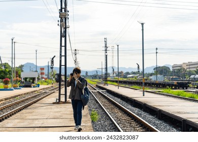 Asian man using digital camera taking picture during walking on railroad track at train station. People enjoy outdoor lifestyle travel countryside by public transportation on summer holiday vacation.