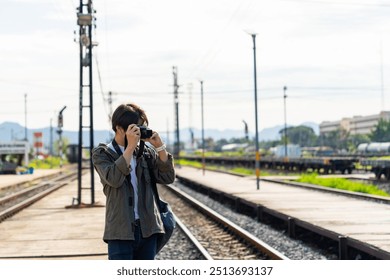 Asian man using digital camera taking picture during walking on railroad track at train station. People enjoy outdoor lifestyle travel countryside by public transportation on summer holiday vacation.