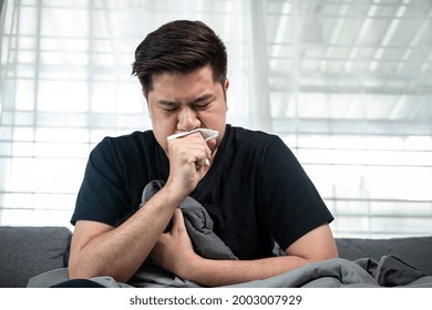 Asian Man Using Tissue In Hand Cover Your Mouth When Cough And Sneezing While Feeling Sick, High Fever And Sitting With Blanket On Couch At Home