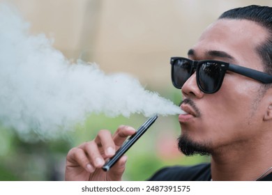 Asian man vaping with an electronic cigarette vape device concept. Smoking with an E-cigarette blowing smoke from mouth, and enjoying outdoors in summer, modern alternative to tobacco addictive habit.