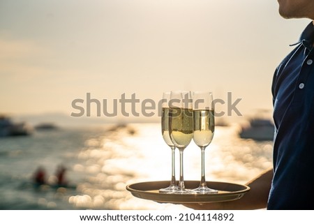 Asian man waiter holding champagne glass on the tray serving to group of passenger tourist travel on luxury catamaran boat yacht sailing in the ocean at summer sunset on beach holiday vacation trip.