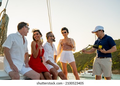 Asian man waiter serving champagne to tourist while catamaran boat sailing at summer sunset. Happy man and woman friends relax and enjoy luxury outdoor lifestyle sail yacht on tropical travel vacation